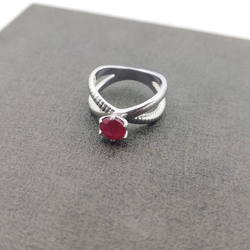 May include: A silver ring with a crisscross design. The ring features a round, bright red gemstone at the center. The band has a textured pattern on one side. The ring is set against a gray background.