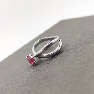 May include: A silver ring with a red gemstone. The ring has a unique design with a band that crosses over itself. The gemstone is round and set in a prong setting. The ring is displayed on a gray surface.