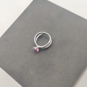 May include: A silver ring with a split band design, featuring a round, red gemstone. The ring is set on a gray surface, showcasing its elegant and modern style. The gemstone is the focal point, adding a pop of color to the silver band.