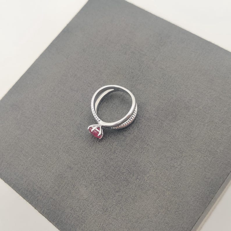 May include: A silver ring with a split band design, featuring a round, red gemstone. The ring is set on a gray surface, showcasing its elegant and modern style. The gemstone is the focal point, adding a pop of color to the silver band.