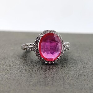 Könnte beinhalten: Ein silberner Ring mit einem großen, ovalen, magentafarbenen Edelstein, umgeben von einem Kranz aus kleineren, klaren Steinen. Der Ring ist ebenfalls mit kleineren, klaren Steinen besetzt, was einen funkelnden Effekt erzeugt. Der Ring ist vor grauem Hintergrund platziert.