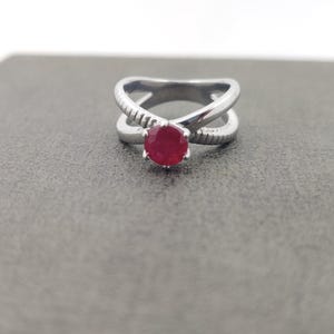 May include: A silver ring with a crisscross design and a round, red gemstone. The ring's band has a textured pattern on the crossing sections. The gemstone is set in a six-prong setting, showcasing its vibrant color.