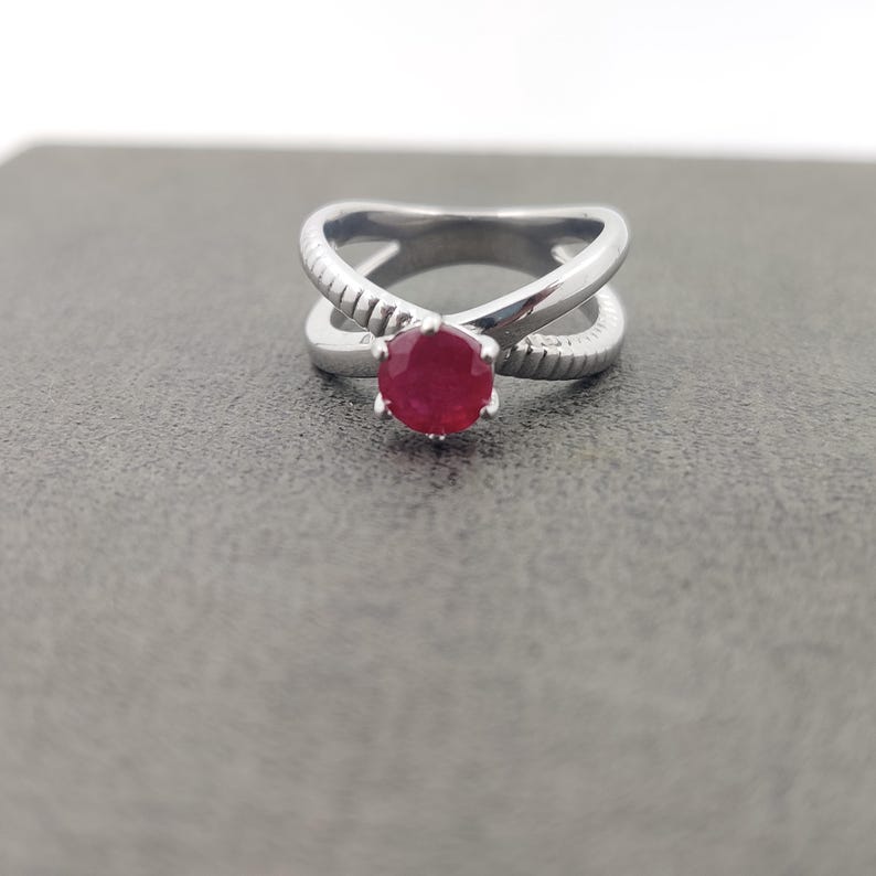 May include: A silver ring with a crisscross design and a round, red gemstone. The ring's band has a textured pattern on the crossing sections. The gemstone is set in a six-prong setting, showcasing its vibrant color.