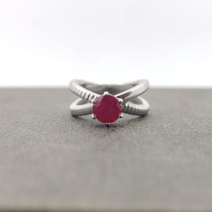 May include: A silver ring with a crisscross design and a large, round, red gemstone. The band has textured detailing on the outer edges. The ring is set against a gray background.