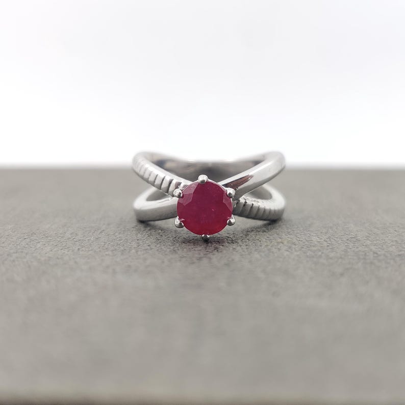 May include: A silver ring with a crisscross design and a large, round, red gemstone. The band has textured detailing on the outer edges. The ring is set against a gray background.