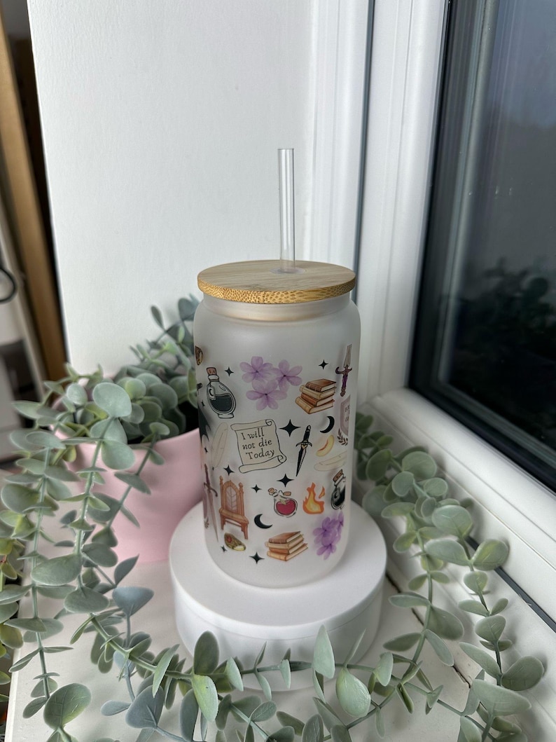 K&ouml;nnte beinhalten: Ein mattierter Glasbecher mit Bambusdeckel und Strohhalm. Der Becher ist mit verschiedenen Stickern im Hexenstil verziert, darunter ein Halbmond, ein Kessel, ein Buch und ein Dolch. Der Text "Ich werde heute nicht sterben" ist auf den Becher gedruckt.