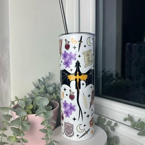 Op de afbeelding: Een witte roestvrijstalen tumbler met een zwarte draak, paarse bloemen, sterren, manen en andere magische symbolen. De tumbler heeft een zwart deksel en een zwarte rietje. De tekst "Fly or Die" is zichtbaar op de tumbler.