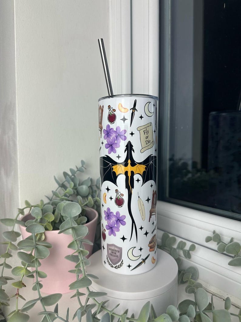 Op de afbeelding: Een witte roestvrijstalen tumbler met een zwarte draak, paarse bloemen, sterren, manen en andere magische symbolen. De tumbler heeft een zwart deksel en een zwarte rietje. De tekst "Fly or Die" is zichtbaar op de tumbler.