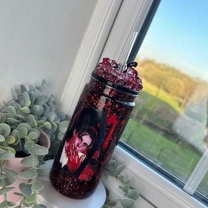 May include: A red glitter tumbler with a black lid and a clear straw. The tumbler features a graphic of a person with red accents. The tumbler is decorated with red glitter and a black spiderweb design.