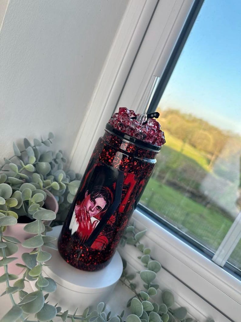 May include: A red glitter tumbler with a black lid and a clear straw. The tumbler features a graphic of a person with red accents. The tumbler is decorated with red glitter and a black spiderweb design.