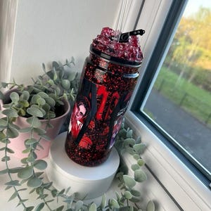 May include: A red glitter tumbler with a black lid and clear straw. The tumbler features a graphic with red and white text and images. The tumbler is on a white pedestal with greenery in the background.