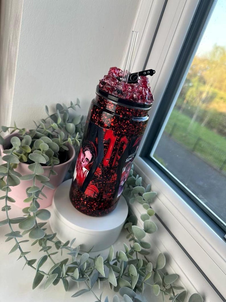 May include: A red glitter tumbler with a black lid and clear straw. The tumbler features a graphic with red and white text and images. The tumbler is on a white pedestal with greenery in the background.