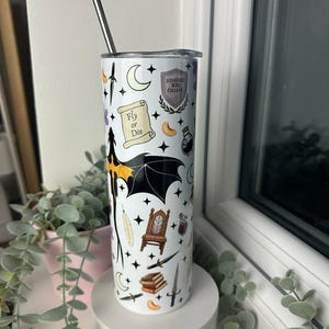 Op de afbeelding: Een witte tumbler met een zwart-wit ontwerp met een draak, een kroon, een veer, een boek, een dolk, een flesje met een toverdrank, een halve maan, sterren en de tekst "Fly or Die" en "Hogwarts Hall College".