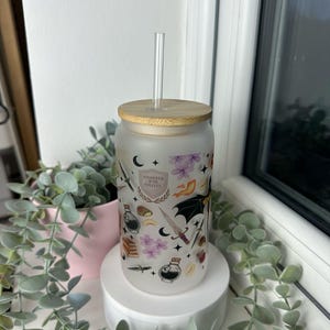 K&ouml;nnte beinhalten: Eine mattierte, wiederverwendbare Wasserflasche aus Glas mit Bambusdeckel und einem klaren Strohhalm. Die Flasche hat ein fantasievolles Design mit einer schwarzen Fledermaus, einem zunehmenden Mond, einem Schwert, einer Zaubertrankflasche und dem Text "Hogwarts War College".