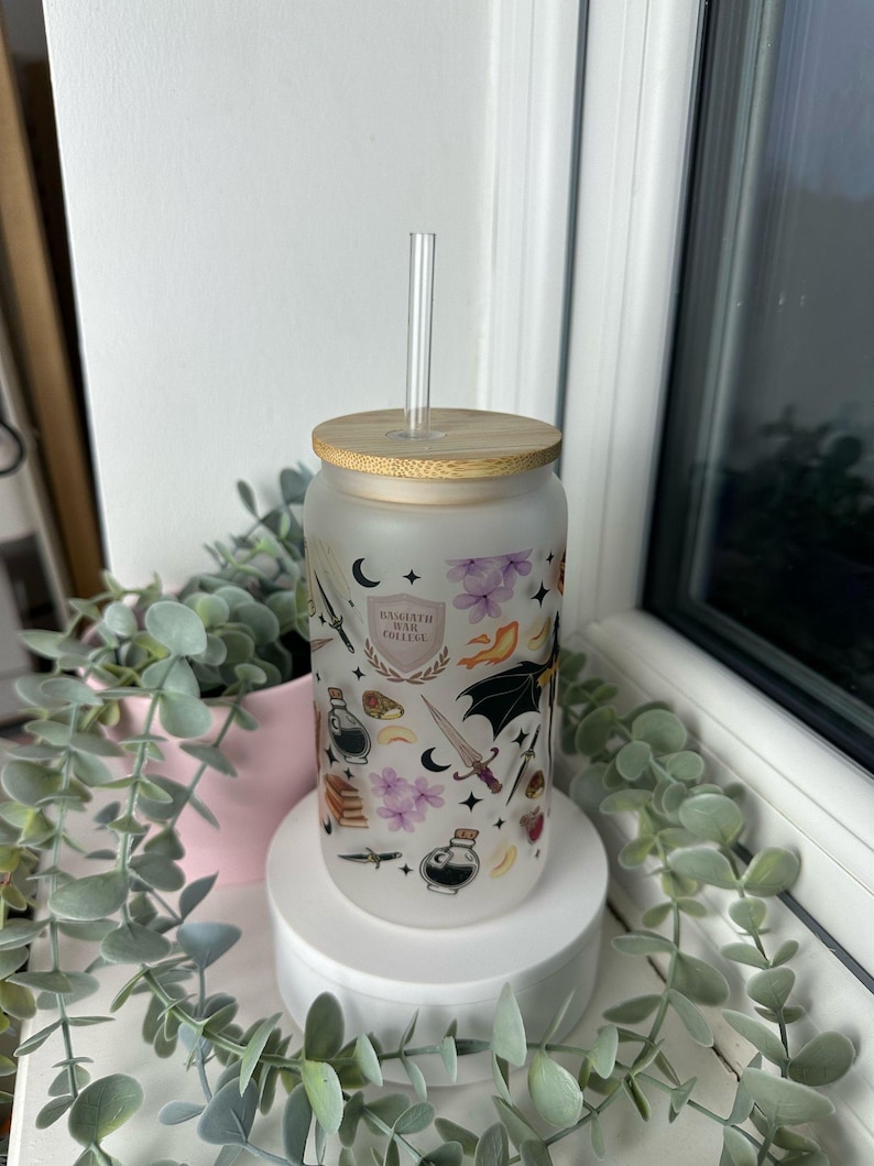 K&ouml;nnte beinhalten: Eine mattierte, wiederverwendbare Wasserflasche aus Glas mit Bambusdeckel und einem klaren Strohhalm. Die Flasche hat ein fantasievolles Design mit einer schwarzen Fledermaus, einem zunehmenden Mond, einem Schwert, einer Zaubertrankflasche und dem Text "Hogwarts War College".