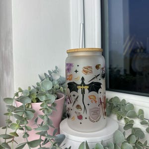 K&ouml;nnte beinhalten: Ein mattierter Glasbecher mit Bambusdeckel und Strohhalm. Der Becher zeigt ein Drachenmotiv mit verschiedenen anderen Illustrationen, darunter B&uuml;cher, Sterne und eine Zaubertrankflasche. Der Text auf dem Becher lautet "Imagination is our collective", "power".
