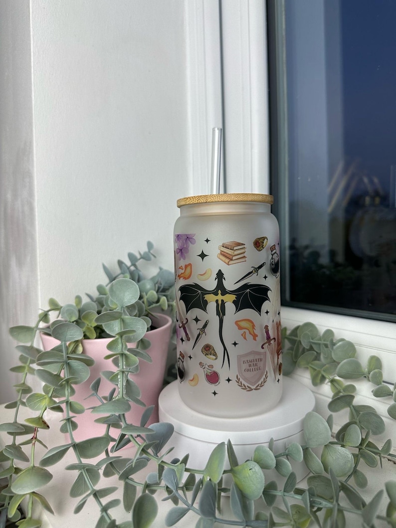 K&ouml;nnte beinhalten: Ein mattierter Glasbecher mit Bambusdeckel und Strohhalm. Der Becher zeigt ein Drachenmotiv mit verschiedenen anderen Illustrationen, darunter B&uuml;cher, Sterne und eine Zaubertrankflasche. Der Text auf dem Becher lautet "Imagination is our collective", "power".