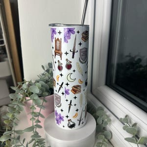Op de afbeelding: Een witte roestvrijstalen beker met een paars, zwart en gouden ontwerp. Het ontwerp bevat verschillende illustraties, waaronder een troon, een pen, een dolk en een boek. De tekst op de beker luidt "Ravenclaw House".