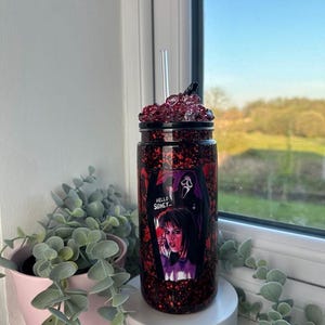 May include: A glitter-covered tumbler with a black lid and a clear straw. The tumbler features a graphic from the movie Scream, with the text "Hello Sidney...". The cup is filled with red glitter and is sitting on a white surface.