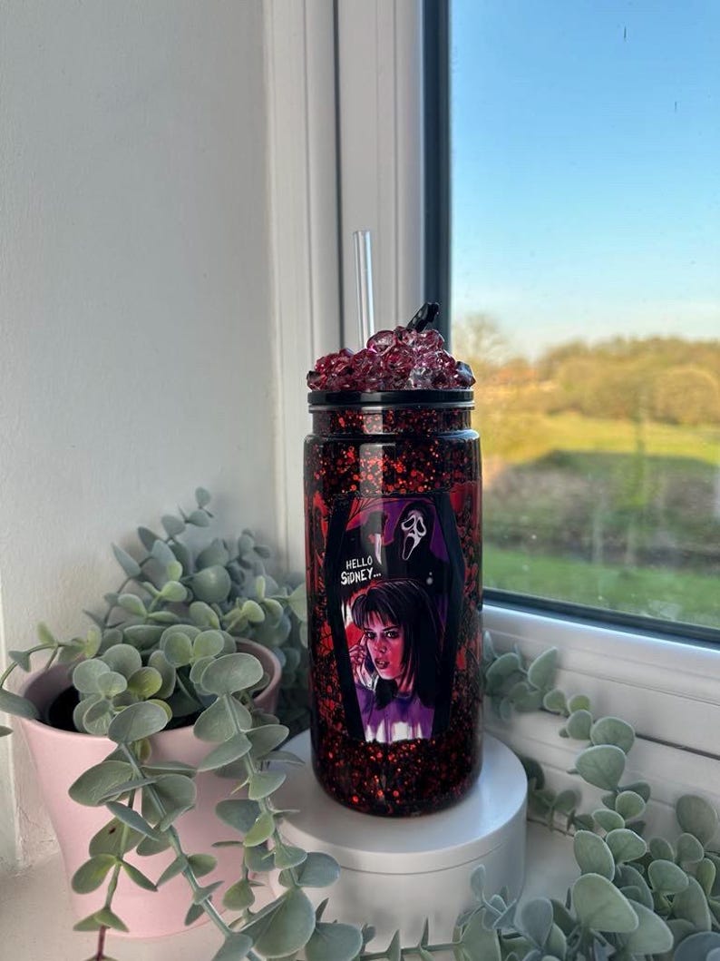 May include: A glitter-covered tumbler with a black lid and a clear straw. The tumbler features a graphic from the movie Scream, with the text "Hello Sidney...". The cup is filled with red glitter and is sitting on a white surface.