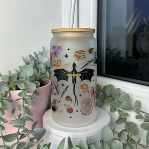 K&ouml;nnte beinhalten: Ein mattierter Glasbecher mit Bambusdeckel und Strohhalm. Der Becher hat ein schwarz-wei&szlig;es Design mit einem Drachen, B&uuml;chern, Schwertern und anderen Fantasy-Motiven. Der Text "Hufflepuff House College" ist auf den Becher gedruckt.
