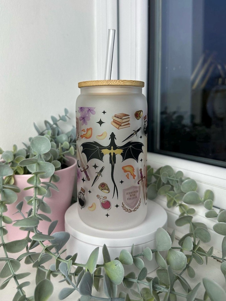 K&ouml;nnte beinhalten: Ein mattierter Glasbecher mit Bambusdeckel und Strohhalm. Der Becher hat ein schwarz-wei&szlig;es Design mit einem Drachen, B&uuml;chern, Schwertern und anderen Fantasy-Motiven. Der Text "Hufflepuff House College" ist auf den Becher gedruckt.