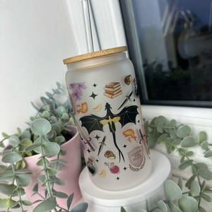 K&ouml;nnte beinhalten: Ein mattierter Glasbecher mit Bambusdeckel und Strohhalm. Der Becher ist mit einem Design aus einem Drachen, B&uuml;chern, Schwertern und anderen Fantasy-Motiven verziert. Der Text "DANGEROUSLY IN LOVE WITH COLLEGE" ist auf den Becher gedruckt.
