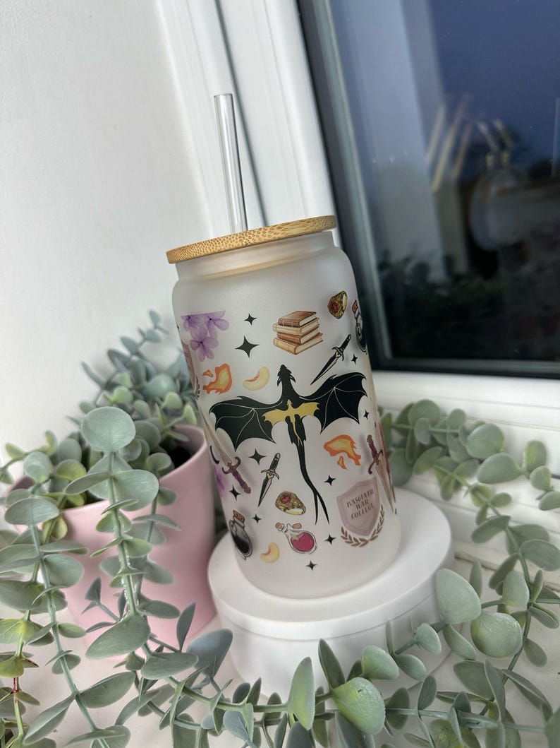 K&ouml;nnte beinhalten: Ein mattierter Glasbecher mit Bambusdeckel und Strohhalm. Der Becher ist mit einem Design aus einem Drachen, B&uuml;chern, Schwertern und anderen Fantasy-Motiven verziert. Der Text "DANGEROUSLY IN LOVE WITH COLLEGE" ist auf den Becher gedruckt.