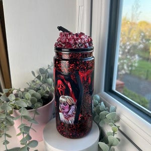 May include: A red glitter tumbler with a black lid and a clear straw. The tumbler features a graphic of a woman and a ghost face from the movie Scream. The top of the tumbler is decorated with red crystals.