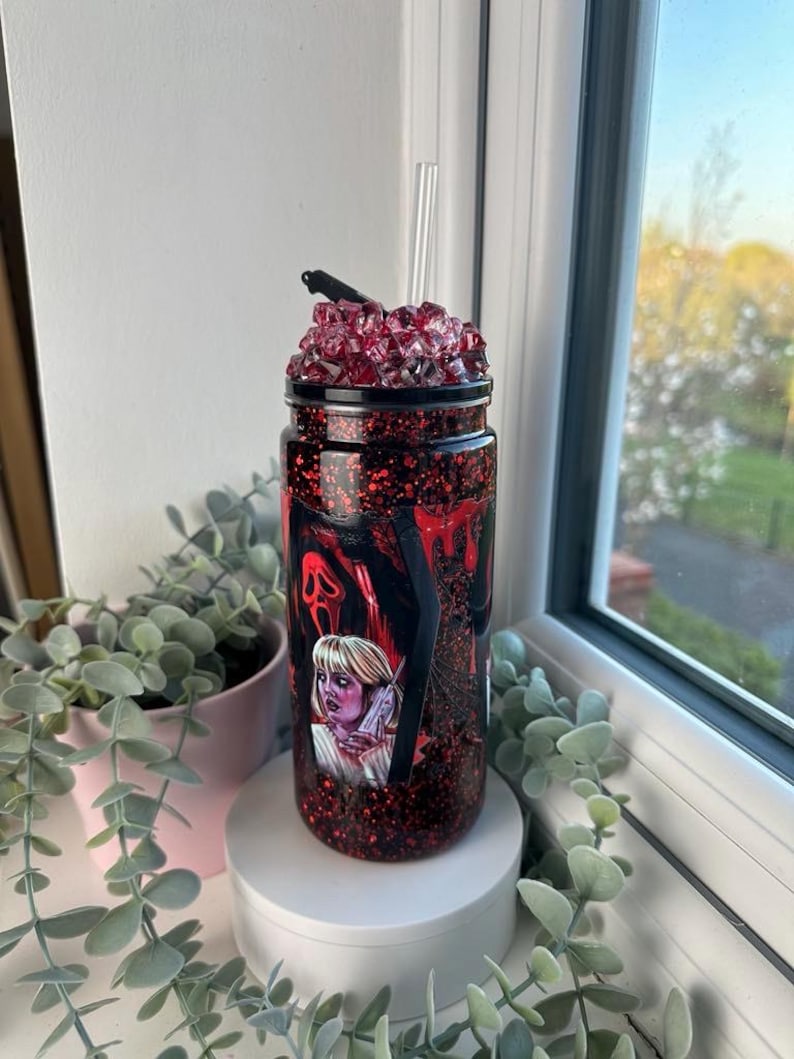 May include: A red glitter tumbler with a black lid and a clear straw. The tumbler features a graphic of a woman and a ghost face from the movie Scream. The top of the tumbler is decorated with red crystals.