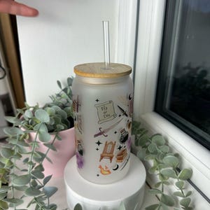 K&ouml;nnte beinhalten: Ein gefrosteter Glasbecher mit Bambusdeckel und Strohhalm. Der Becher hat ein Design mit verschiedenen Illustrationen, darunter ein Schwert, eine Krone, ein Buch und eine Zaubertrankflasche. Der Text "Fly or Die" ist auf einer Schriftrolle im Design gedruckt.