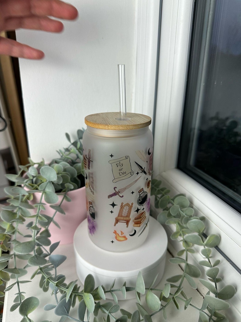 K&ouml;nnte beinhalten: Ein gefrosteter Glasbecher mit Bambusdeckel und Strohhalm. Der Becher hat ein Design mit verschiedenen Illustrationen, darunter ein Schwert, eine Krone, ein Buch und eine Zaubertrankflasche. Der Text "Fly or Die" ist auf einer Schriftrolle im Design gedruckt.