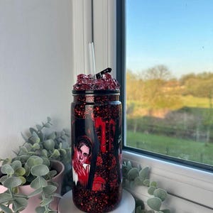 May include: A red glitter tumbler with a black lid and a clear straw. The tumbler features a graphic of a person in a red and black design. The top of the tumbler is filled with red crystals. The tumbler is sitting on a white surface.