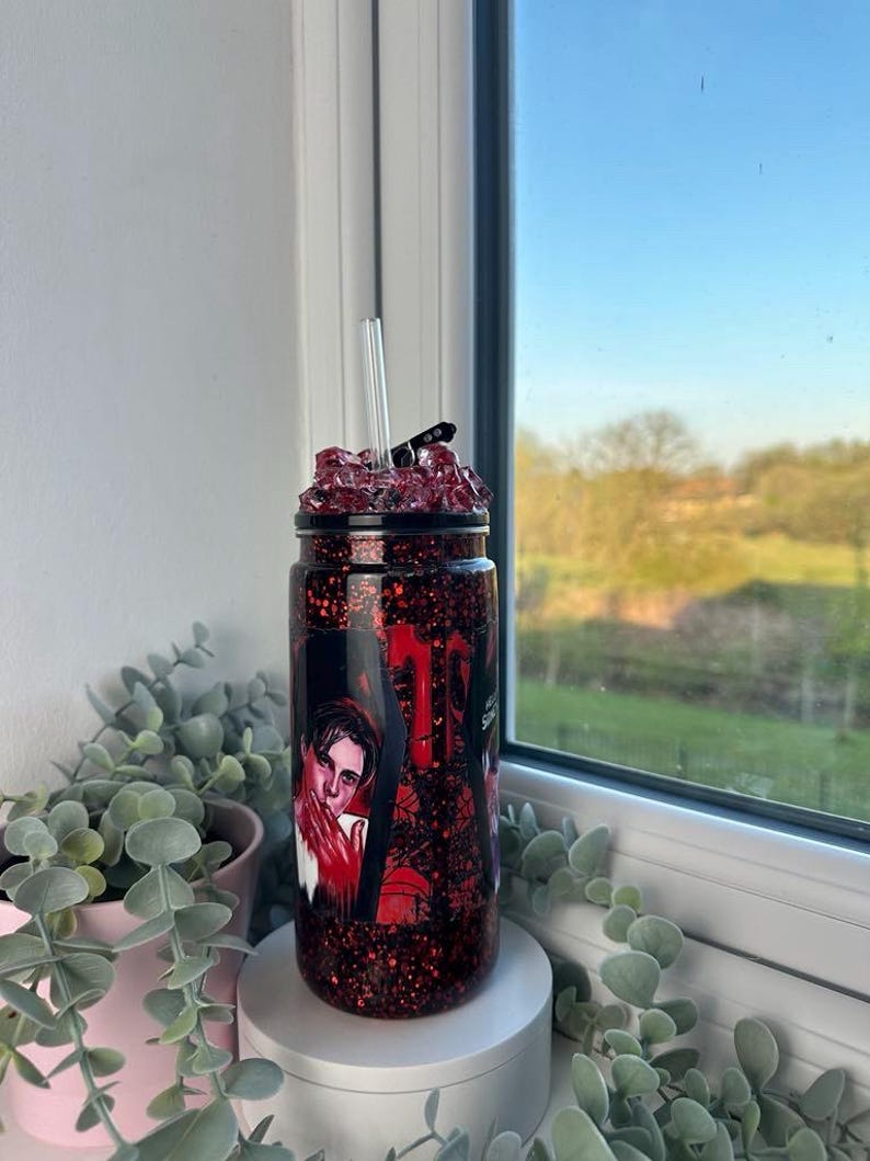 May include: A red glitter tumbler with a black lid and a clear straw. The tumbler features a graphic of a person in a red and black design. The top of the tumbler is filled with red crystals. The tumbler is sitting on a white surface.