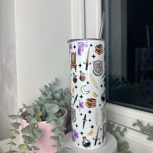 Op de afbeelding: Een witte roestvrijstalen beker met een zwart-wit patroon van verschillende magische voorwerpen, waaronder boeken, veren, drankjes, dolken en een wapenschild met de tekst "Hogwarts School voor Hekserij en Toverkunst".