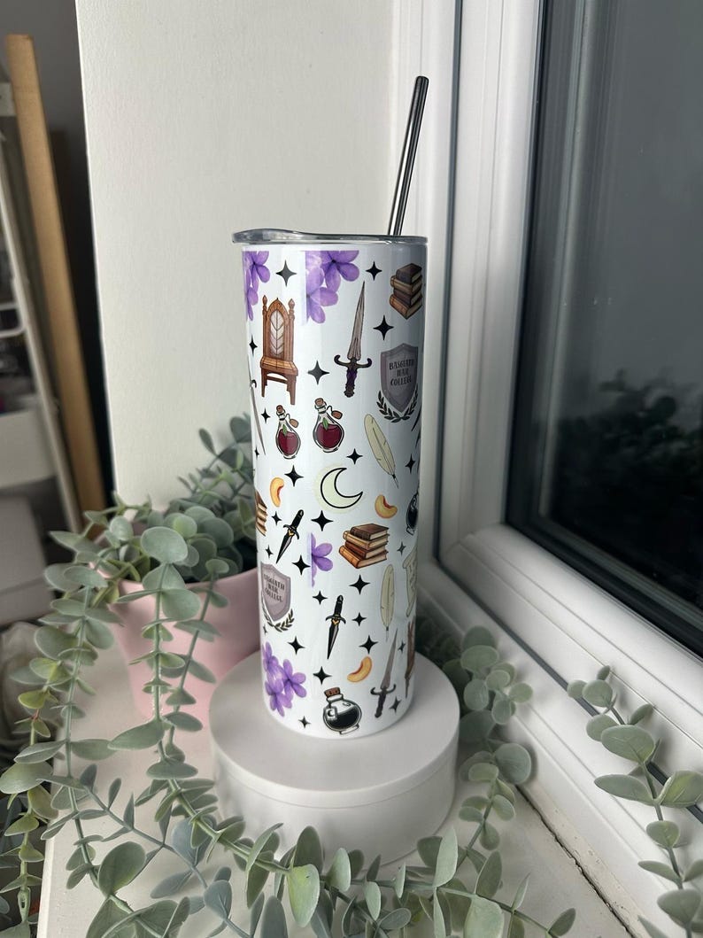 Op de afbeelding: Een witte roestvrijstalen beker met een paars, zwart en gouden ontwerp. De beker heeft verschillende illustraties, waaronder een troon, een pen, een dolk en een boek. De tekst "Ravenclaw House" is zichtbaar op de beker.