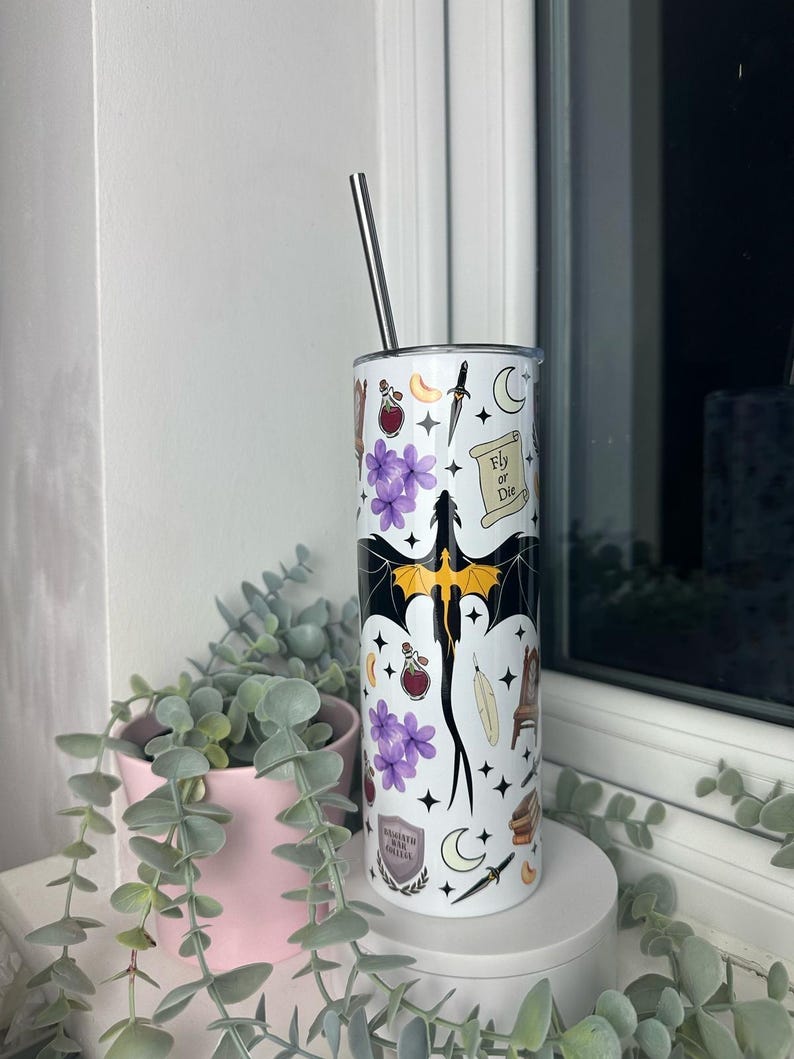 Op de afbeelding: Een witte roestvrijstalen tumbler met een zwart en geel drakenontwerp. De tumbler is bedekt met verschillende stickers, waaronder een halve maan, een veer, een flesje met een toverdrank, een dolk en een rol die "Fly or Die" zegt.