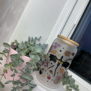 K&ouml;nnte beinhalten: Ein mattierter Glasbecher mit Bambusdeckel und einem klaren Strohhalm. Der Becher ist mit einem schwarzen Drachen, Sternen und verschiedenen anderen Fantasy-Motiven verziert. Der Text "Always be kind to witches" ist auf den Becher gedruckt.