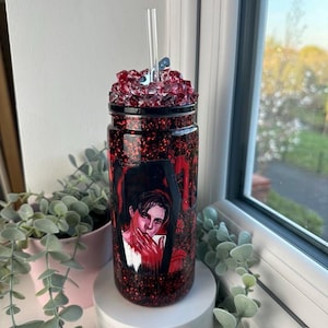 May include: A red and black glitter tumbler with a portrait design. The tumbler has a clear straw and is topped with red crystal-like embellishments. The tumbler is displayed on a white pedestal. The design has red accents.