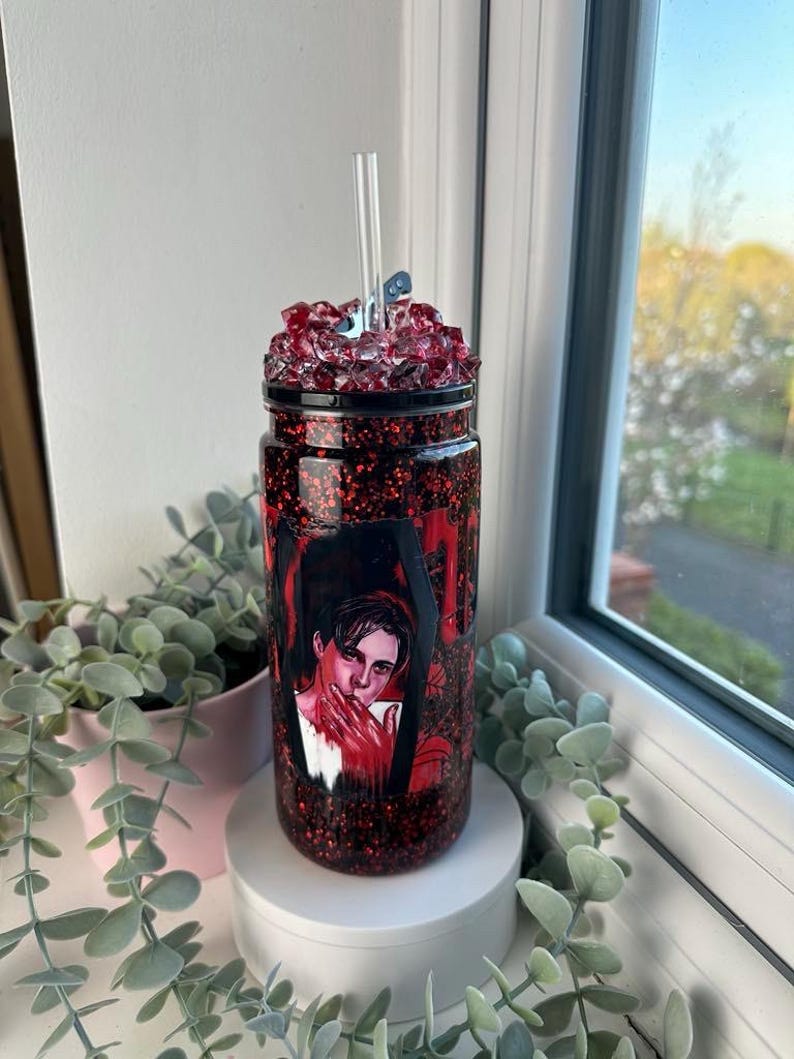 May include: A red and black glitter tumbler with a portrait design. The tumbler has a clear straw and is topped with red crystal-like embellishments. The tumbler is displayed on a white pedestal. The design has red accents.