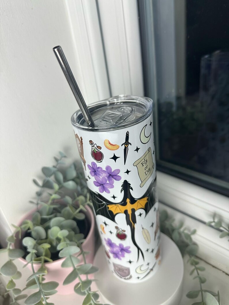 Op de afbeelding: Een witte roestvrijstalen tumbler met een zwarte draak en verschillende magische symbolen, waaronder een flesje met een toverdrank, een dolk, een halve maan en een rol met de tekst "Fly or Die".
