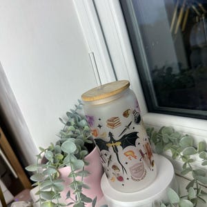 K&ouml;nnte beinhalten: Ein gefrosteter Glasbecher mit Bambusdeckel und Strohhalm. Der Becher zeigt ein fantasievolles Design mit einem Drachen, Sternen, B&uuml;chern und anderen magischen Elementen. Der Text auf dem Becher lautet "Brewed with Magic Coffee".