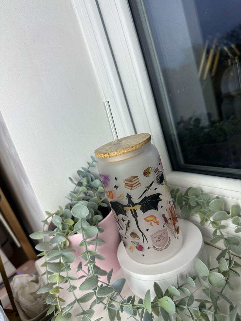 K&ouml;nnte beinhalten: Ein gefrosteter Glasbecher mit Bambusdeckel und Strohhalm. Der Becher zeigt ein fantasievolles Design mit einem Drachen, Sternen, B&uuml;chern und anderen magischen Elementen. Der Text auf dem Becher lautet "Brewed with Magic Coffee".