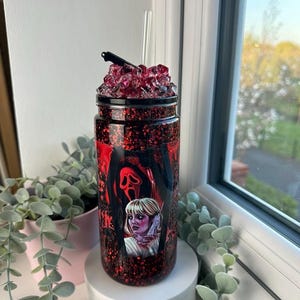 May include: A glittery tumbler with a horror movie theme. The tumbler features a design with a Scream mask and a woman holding a knife. The lid is topped with red crystals and a small knife. A clear straw is inserted into the tumbler.