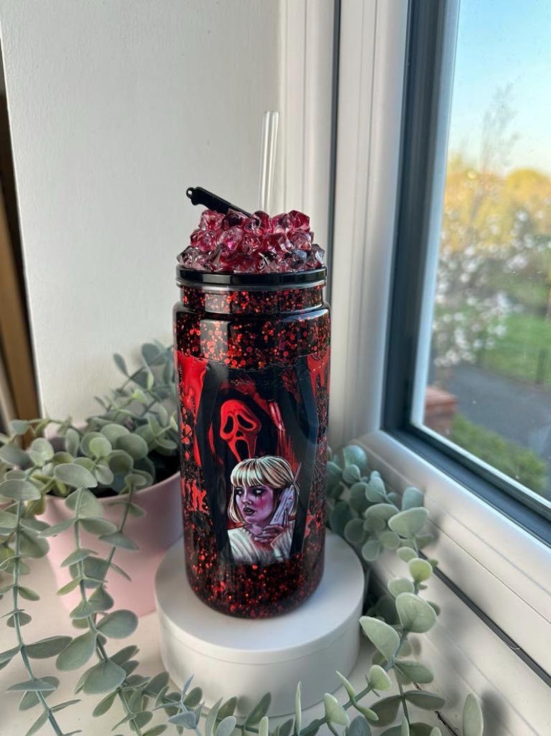 May include: A glittery tumbler with a horror movie theme. The tumbler features a design with a Scream mask and a woman holding a knife. The lid is topped with red crystals and a small knife. A clear straw is inserted into the tumbler.
