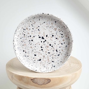 Op de afbeelding: Een witte keramische schaal met een terrazzo-patroon in zwart, bruin en beige. De schaal staat op een houten voetstuk.
