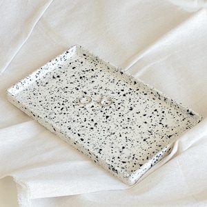 Könnte beinhalten: Ein weißes rechteckiges Tablett mit schwarzen Flecken, perfekt zum Aufbewahren von Schmuck oder anderen kleinen Gegenständen. Das Tablett liegt auf einer weißen Stoffoberfläche.