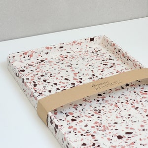 May include: A rectangular white tray with a pink and brown terrazzo pattern. The tray has a brown paper label with the text "douce maison" on it.
