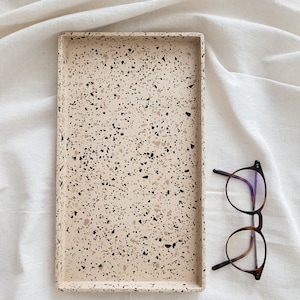 Könnte beinhalten: Ein rechteckiges Tablett mit einem gesprenkelten beige, schwarzen und braunen Terrazzo-Muster. Das Tablett befindet sich auf einem weißen Leinenhintergrund.