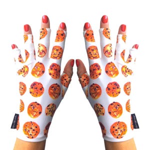 May include: White gloves with a repeating pattern of chocolate chip cookies. The gloves have a black tag on the wrist area. The fingers are exposed, and the nails are painted red.